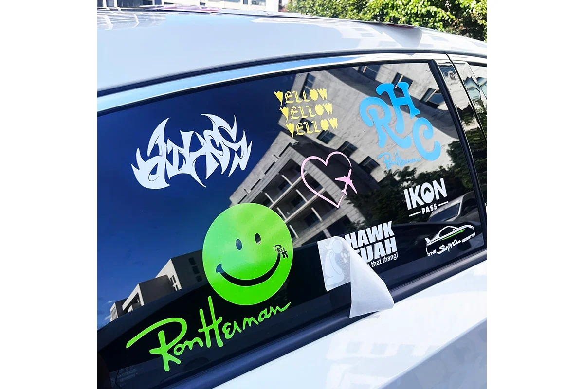 A car side window displaying printed logos, lettering and a green smiley face.