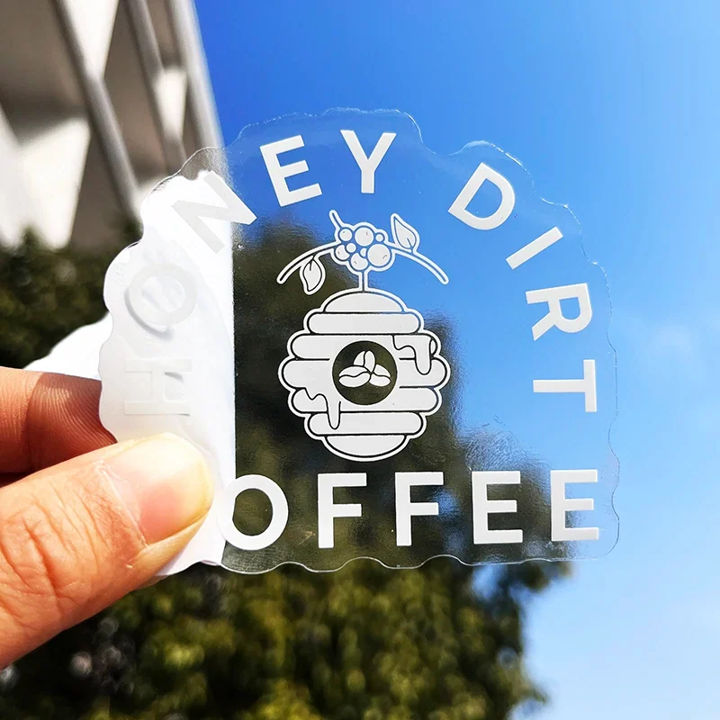 A clear custom-outline coffee logo sticker held against a blue background