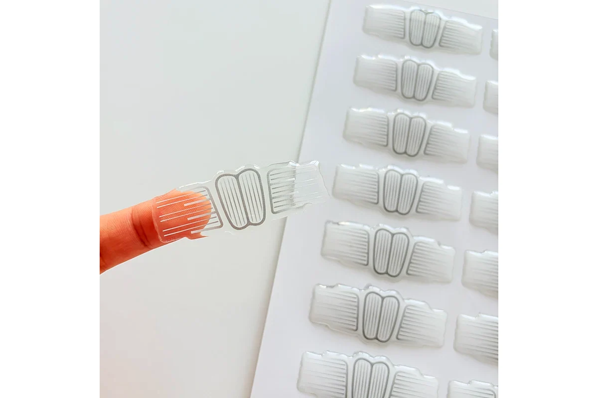 A clear shaped domed label lifted on a fingertip beside matching labels on a sheet.