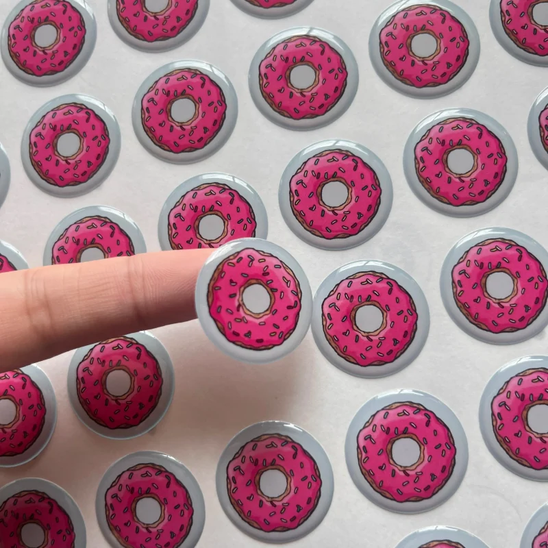 A hand holding a pink donut design epoxy sticker above a printed sheet.
