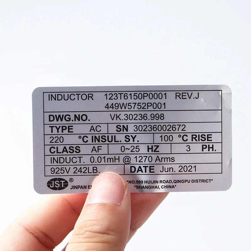 A hand holding a silver industrial equipment label printed with identification specifications.