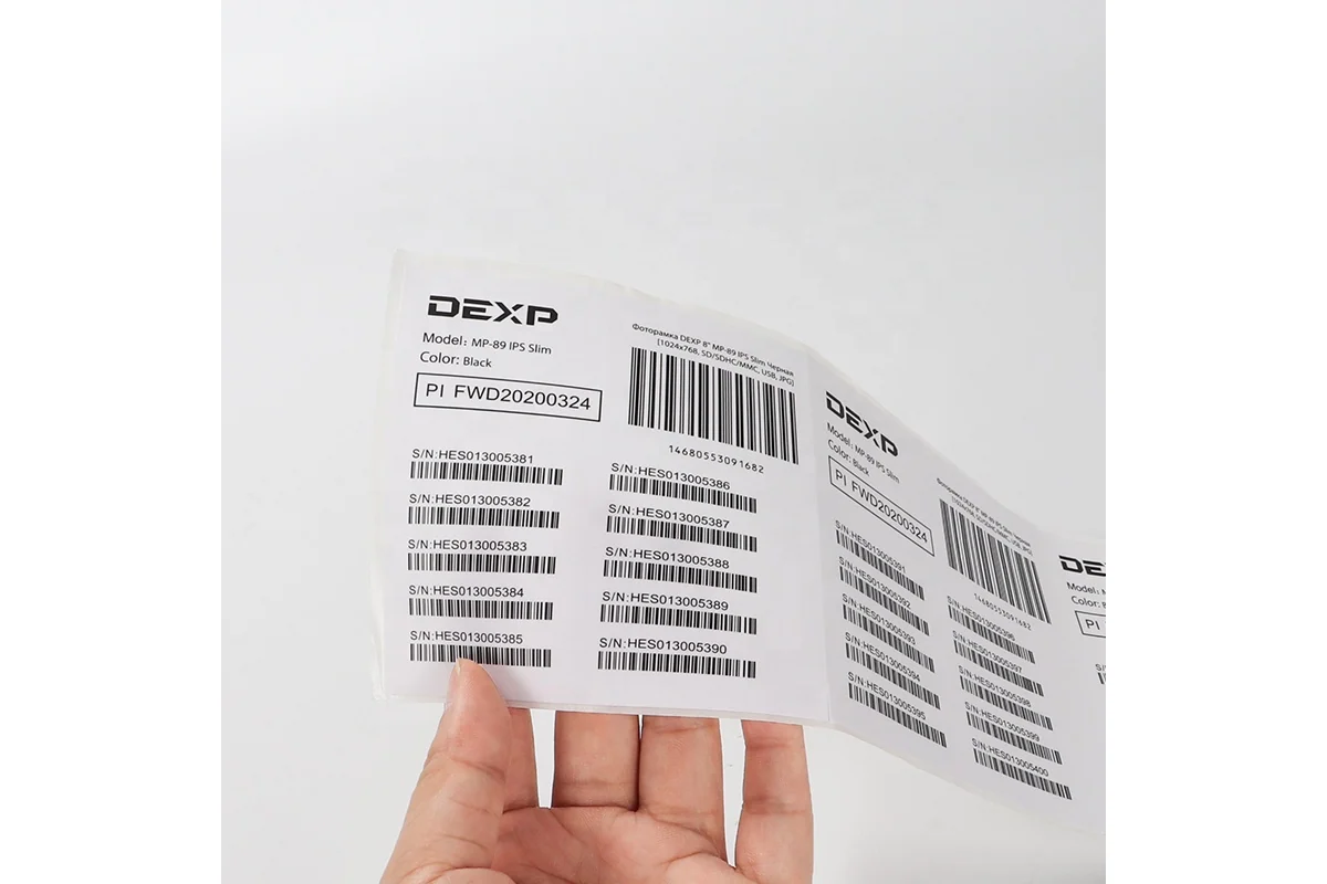 A hand holding a sheet of white equipment labels printed with serial numbers and barcodes.