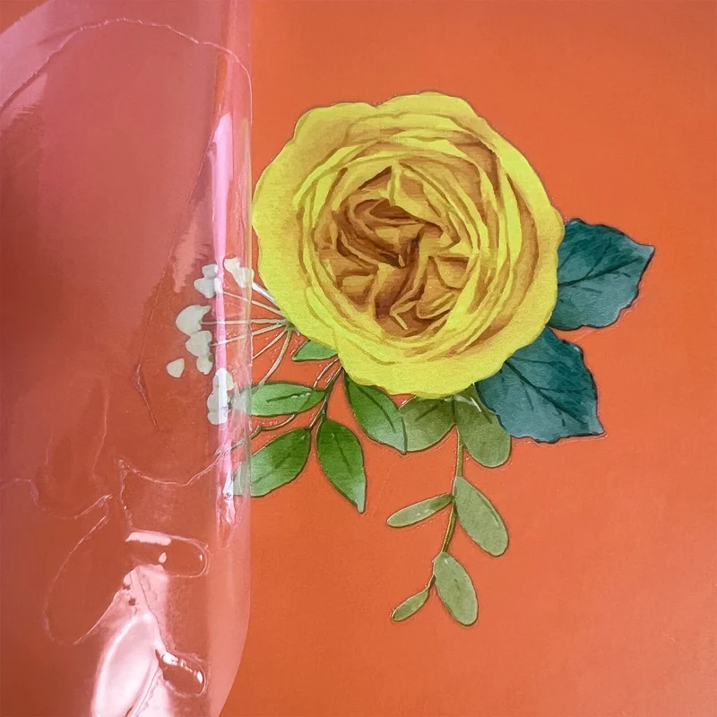 A yellow rose and green leaves transferred to an orange surface as clear film is lifted.